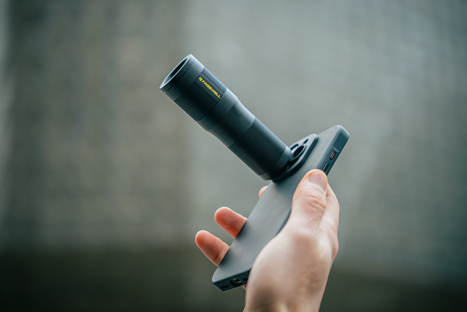 Hand holding an iPhone equipped with a Freewell 6x telephoto lens against a blurred gray background.