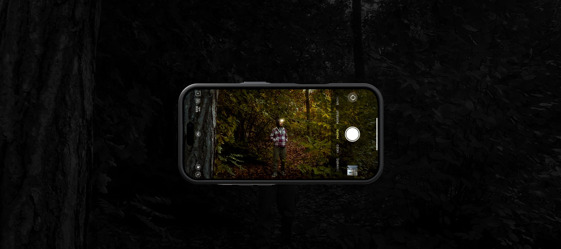 A person standing in a forest with autumn leaves, captured on an iPhone camera screen showing a photo taken in a dimly lit environment, with the phone's camera app interface visible without using Freewell 1.33x anamorphic lens.